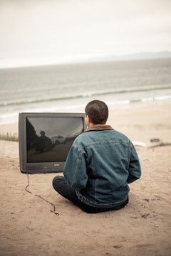 A person sitting on a sandy beach facing a large television screen. The ocean and horizon are visible in the background, with overcast skies creating a muted atmosphere. The individual wears a denim jacket and is kneeling near the unplugged TV.