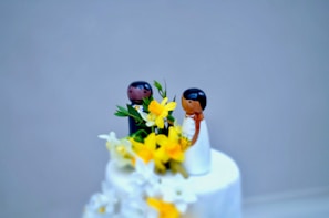 A wedding cake topped with two wooden figurines depicting a couple, surrounded by vibrant yellow flowers and green leaves. The cake's surface is smooth and white, creating a crisp and elegant appearance.