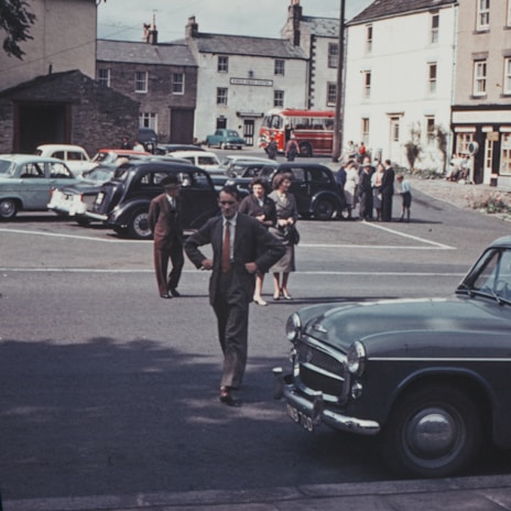 A vintage street scene features several parked cars with a classic design. People, dressed in mid-20th-century attire, stroll or stand nearby. A red bus is visible in the background, and the buildings have a quaint, old-fashioned architectural style.