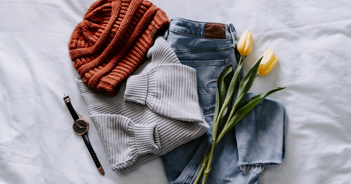 A styled flat lay of clothing items including a knit hat, ribbed sweater, jeans, and accessories on white fabric