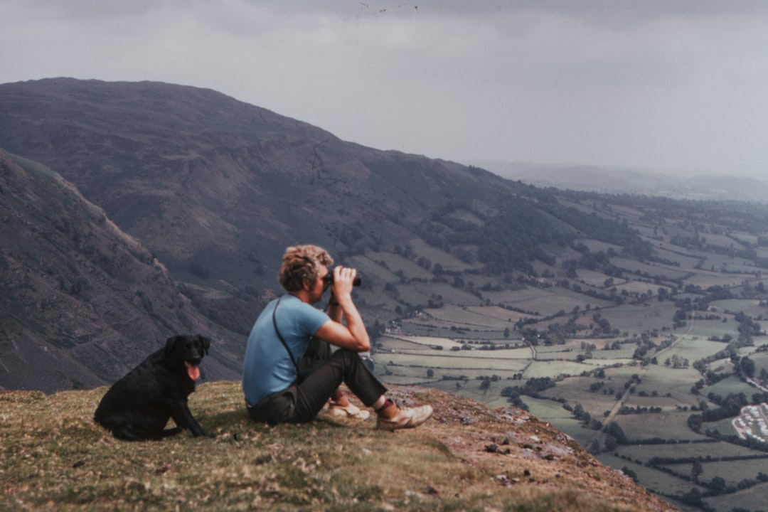 man with binoculars sitting on top of hill beside black dog