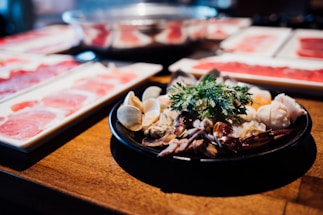A black plate is filled with a variety of seafood, including clams, crab claws, and possibly shrimp. A garnish of fresh green herbs is placed on top. In the background, there are rectangular white plates with thinly sliced red meat arranged in rows. A wooden table provides a warm, rustic backdrop.