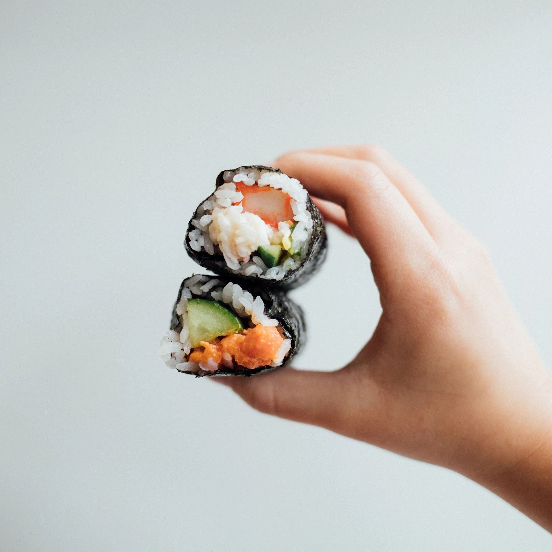 A hand holding two cut maki pieces filled with crab, cucumber and salmon
