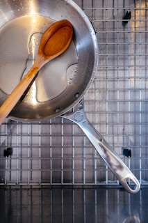 A stainless steel cooking pan with water droplets inside is placed on a metal dish rack. A wooden spoon rests inside the pan.