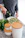 A person in a grey sweater is pouring olive oil over sliced zucchini in a white baking dish. Nearby, there are fresh green herbs and stacked containers with wooden lids on a marble countertop.