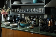 A kitchen countertop featuring a variety of items including two bowls, a blue pot, and a bottle of wine. There are several glass jars and bottles, likely containing oils and vinegars. A bouquet of pink flowers is placed next to a blender. Shelving above the counter holds stacked plates and glasses.
