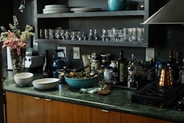 A kitchen countertop featuring a variety of items including two bowls, a blue pot, and a bottle of wine. There are several glass jars and bottles, likely containing oils and vinegars. A bouquet of pink flowers is placed next to a blender. Shelving above the counter holds stacked plates and glasses.