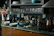 A kitchen countertop featuring a variety of items including two bowls, a blue pot, and a bottle of wine. There are several glass jars and bottles, likely containing oils and vinegars. A bouquet of pink flowers is placed next to a blender. Shelving above the counter holds stacked plates and glasses.