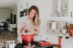 A smiling woman holding a bright lines kitchen utensil in a cozy, sunlit kitchen.
