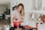 A smiling woman holding a weight scale in a bright kitchen.