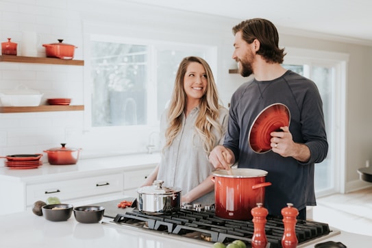 A couple is in a bright, modern kitchen with white cabinetry and large windows providing ample natural light. The man is holding a red pot lid and appears to be cooking, while the woman stands next to him, smiling. Various kitchen items, including red pots and black bowls, are arranged on the countertop and stove.