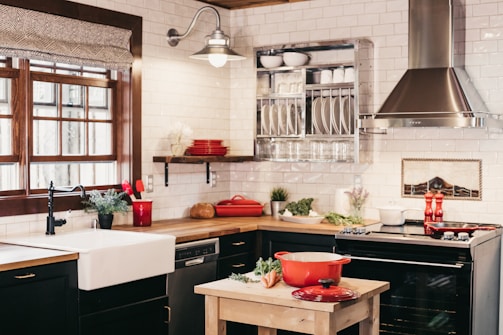 A warm and inviting kitchen with rustic elements includes white subway tiles and a large farmhouse sink. A wooden countertop enhances the cozy feel, complemented by dark cabinetry and stainless steel appliances. A red pot sits atop a wooden table, surrounded by fresh vegetables, hinting at cooking activity. Various herbs and potted plants add a touch of greenery, while red utensils and dishware provide pops of color throughout the room.
