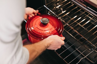A person is placing a red cast iron pot into an open oven, highlighting the pot's vibrant color and glossy surface against the oven's metallic racks.
