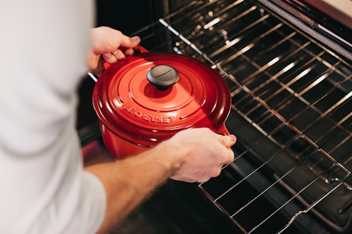 A person is placing a red cast iron pot into an open oven, highlighting the pot's vibrant color and glossy surface against the oven's metallic racks.