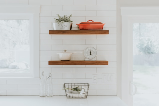 A minimalist kitchen scene featuring white tiled walls and wooden shelves. The top shelf holds a red pot and a decorative plant in a white container labeled 'FLEURS JARDIN.' The lower shelf displays a white saucepan next to a vintage-style kitchen scale. Below the shelves, there are two clear glass bottles and a small basket containing rolled textiles.