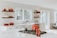 A bright and spacious kitchen features white cabinetry and countertops, with a sleek, modern design. A red pot sits on the stove accompanied by matching red salt and pepper shakers. Wooden shelves display various kitchen items, including more red cookware. Large windows allow natural light to pour in, enhancing the airy atmosphere.