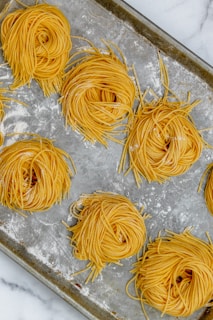 Several bundles of raw, yellow pasta nests arranged evenly on a well-used, textured baking sheet. The sheet has a light dusting of flour, and each pasta nest is neatly coiled, displaying a uniform look in size and shape.