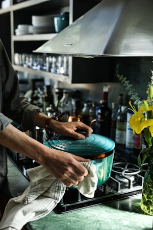 A person is holding a turquoise Dutch oven on a stove. Nearby, various bottles are lined up on a countertop, and a bright yellow flower is visible in a vase. The kitchen includes a stainless steel hood and open shelving with glassware and dishes.