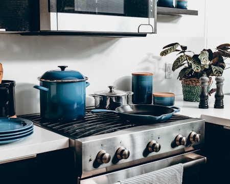 A modern kitchen scene features a stovetop with a blue pot and a pan. Nearby, several containers with wooden lids and a wicker basket containing a plant are visible. The setting suggests a clean and organized kitchen environment.