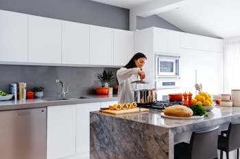 A woman cooking in a modern kitchen with white cabinets and stainless steel appliances. The countertop is filled with various kitchen items including fruits, vegetables, and a loaf of bread. Bright sunlight streams in from a window, illuminating the scene.