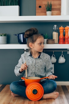 A young girl with brown hair tied up sits cross-legged on a wooden floor, stirring a wooden spoon in an orange pot placed in front of her. She is wearing a light blue, polka-dotted long-sleeve shirt and jeans. Behind her is a modern kitchen setup with white shelves containing potted plants, kitchen containers, and mugs hanging below. The wall is painted in a dark teal color.