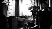 A black and white scene capturing a film set, highlighting a man in profile who appears to be a director or camera operator. He is observing or interacting with a large camera setup, which is positioned on a dolly track. The background features diffused lighting and blurred frames on the walls, suggesting an indoor setting.