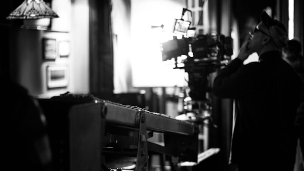 A black and white scene capturing a film set, highlighting a man in profile who appears to be a director or camera operator. He is observing or interacting with a large camera setup, which is positioned on a dolly track. The background features diffused lighting and blurred frames on the walls, suggesting an indoor setting.