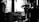 A black and white scene capturing a film set, highlighting a man in profile who appears to be a director or camera operator. He is observing or interacting with a large camera setup, which is positioned on a dolly track. The background features diffused lighting and blurred frames on the walls, suggesting an indoor setting.