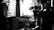 A black and white scene capturing a film set, highlighting a man in profile who appears to be a director or camera operator. He is observing or interacting with a large camera setup, which is positioned on a dolly track. The background features diffused lighting and blurred frames on the walls, suggesting an indoor setting.