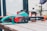 A Bosch electric chainsaw is placed on a wooden table outdoors. The chainsaw is primarily turquoise with black and red accents. Next to it, there's a bottle of chainsaw oil and an orange safety helmet.