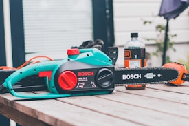 A Bosch electric chainsaw is placed on a wooden table outdoors. The chainsaw is primarily turquoise with black and red accents. Next to it, there's a bottle of chainsaw oil and an orange safety helmet.