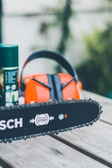 A chainsaw is resting on a wooden surface alongside an orange and black pair of safety earmuffs and a green spray can. The chainsaw bar features a 'Quick Stop' logo.