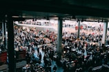 A bustling indoor market with many people browsing various stalls. The vendors have set up tables with an assortment of items for sale, ranging from antiques to everyday goods. The market is held in a large, covered space with natural light coming from above. There are multiple levels visible, with additional shoppers seen on an upper balcony area.