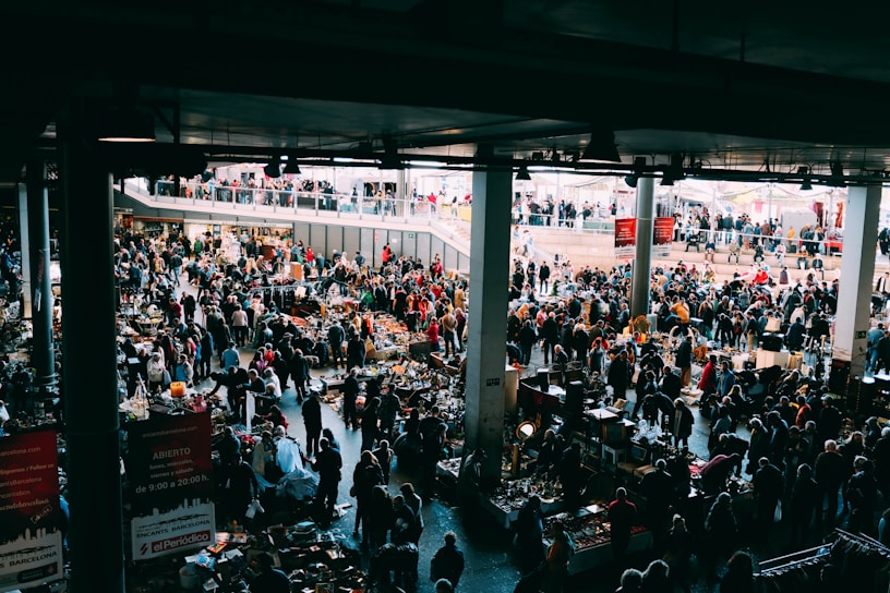 A bustling indoor market with many people browsing various stalls. The vendors have set up tables with an assortment of items for sale, ranging from antiques to everyday goods. The market is held in a large, covered space with natural light coming from above. There are multiple levels visible, with additional shoppers seen on an upper balcony area.