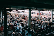 A bustling indoor market with many people browsing various stalls. The vendors have set up tables with an assortment of items for sale, ranging from antiques to everyday goods. The market is held in a large, covered space with natural light coming from above. There are multiple levels visible, with additional shoppers seen on an upper balcony area.