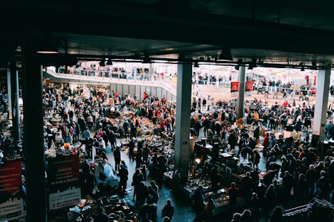 A bustling indoor market with many people browsing various stalls. The vendors have set up tables with an assortment of items for sale, ranging from antiques to everyday goods. The market is held in a large, covered space with natural light coming from above. There are multiple levels visible, with additional shoppers seen on an upper balcony area.