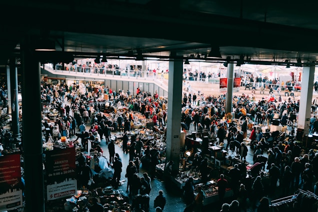 A bustling indoor market with many people browsing various stalls. The vendors have set up tables with an assortment of items for sale, ranging from antiques to everyday goods. The market is held in a large, covered space with natural light coming from above. There are multiple levels visible, with additional shoppers seen on an upper balcony area.