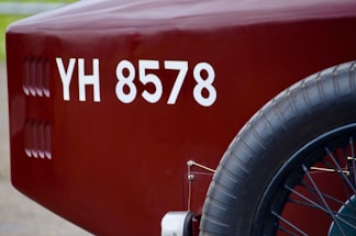 A close-up view of the rear section of a vintage vehicle, displaying a maroon color with a visible license number 'YH 8578'. The image focuses on the spare tire attached to the side and part of the vehicle body.