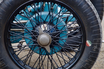 A close-up view of a vintage car wheel with a spoked design. The wheel hub is metallic and has a central cap attached. The tire is branded and features a small round decal with green, white, and red stripes on the side.