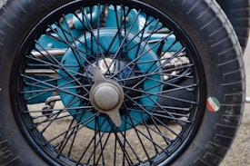 A close-up view of a vintage car wheel with a spoked design. The wheel hub is metallic and has a central cap attached. The tire is branded and features a small round decal with green, white, and red stripes on the side.