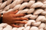 A hand adorned with a black bracelet rests on a chunky, light-colored, braided wool material. The wool appears soft and fluffy.