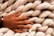 A hand adorned with a black bracelet rests on a chunky, light-colored, braided wool material. The wool appears soft and fluffy.
