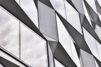 A modern architectural facade featuring angular, geometric shapes with protruding elements. The design includes reflective glass panels showcasing a cloudy sky, creating a dynamic and abstract pattern.