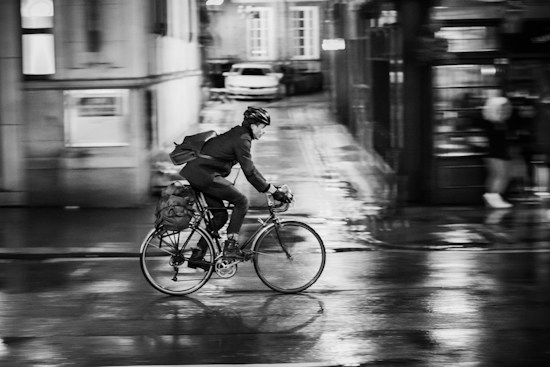 A person is cycling through a wet urban street at night. The cyclist is wearing a helmet and a backpack, and the motion is captured with a blurred effect, emphasizing speed and movement. Reflections of city lights are visible on the slick pavement, suggesting recent rain.
