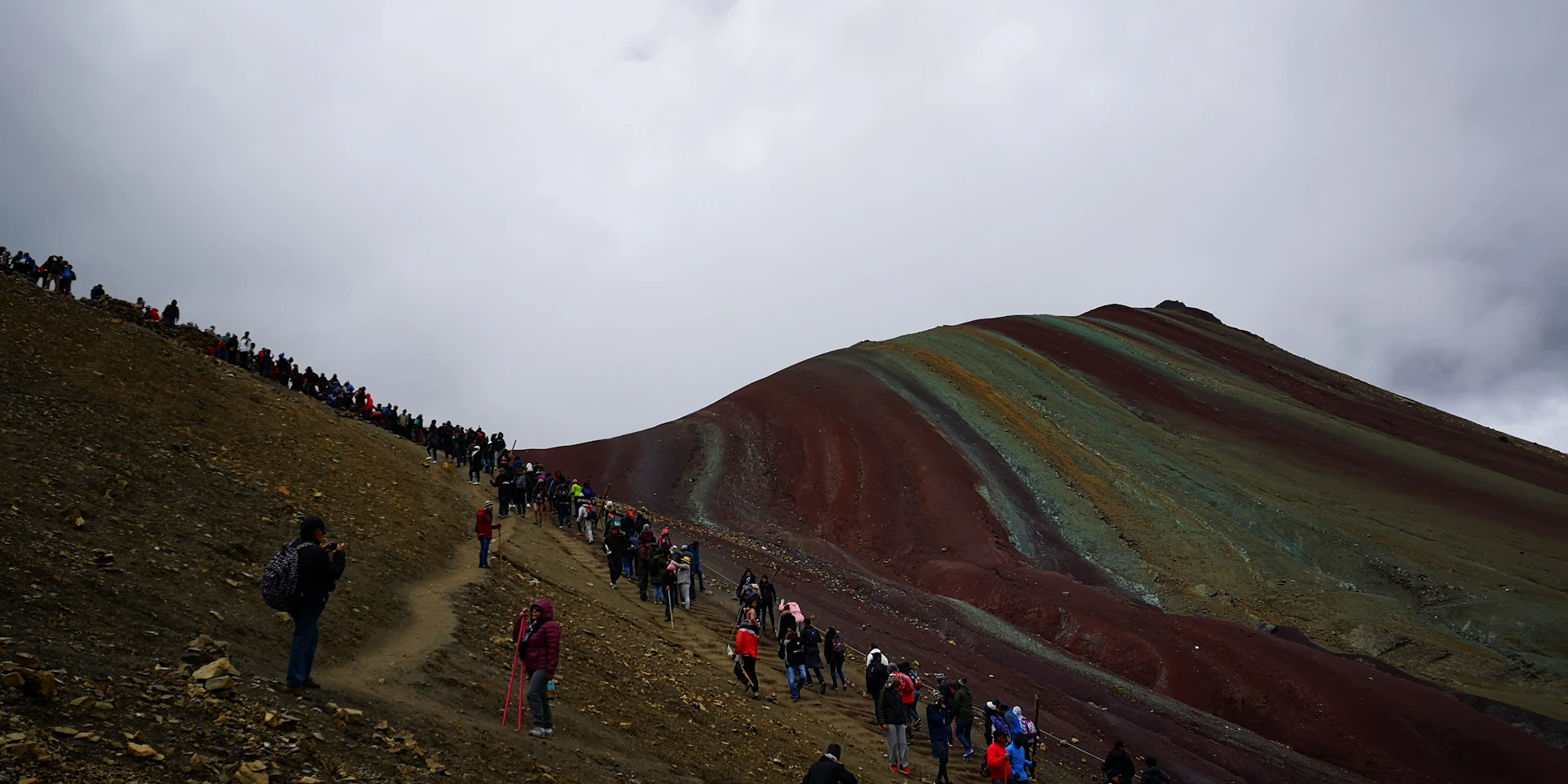 A lively scene of tourists hiking the colorful Rainbow Mountain under a clear blue sky, surrounded by stunning Andean landscapes.