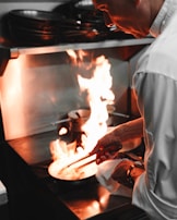 A chef is cooking in a professional kitchen, holding tongs and working over a stovetop with a bright flame. The chef wears a white coat and appears focused on the cooking task.