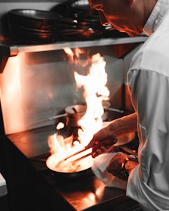 A chef is cooking in a professional kitchen, holding tongs and working over a stovetop with a bright flame. The chef wears a white coat and appears focused on the cooking task.