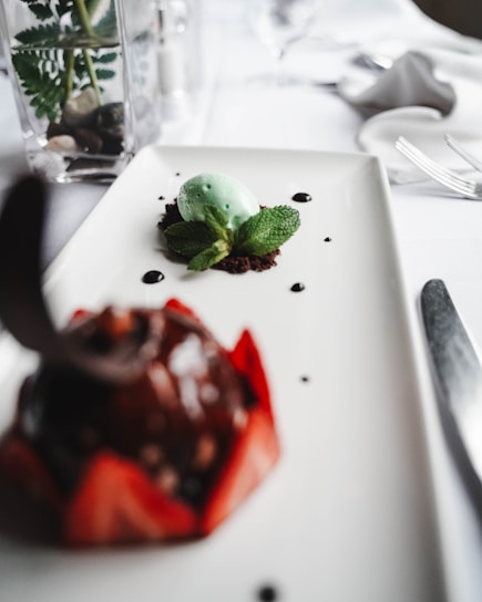 An elegant dessert presentation is displayed on a white rectangular plate. At the foreground, a blurred chocolate dessert with a red decorative base is visible. Behind it, there is a mint green sorbet or mousse placed on a bed of chocolate crumbs, garnished with fresh mint leaves. Small drops of dark syrup or sauce are artfully placed on the plate. In the background, part of a table setting with a knife and a decorative vase with a plant can be seen.