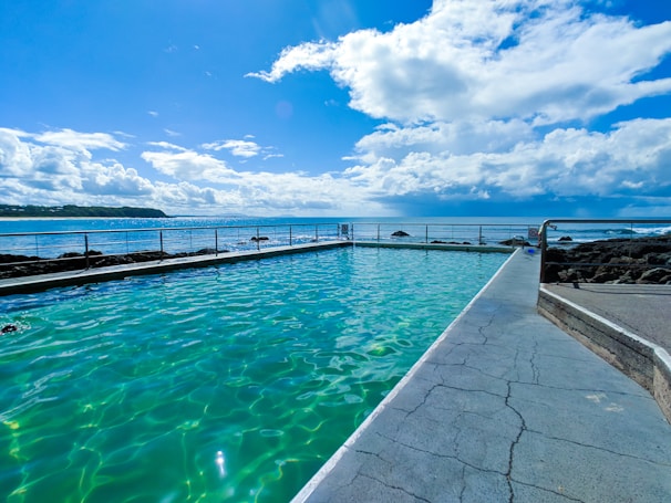 An oceanfront swimming pool with clear turquoise water is surrounded by a concrete deck. The sea is visible in the background under a partially cloudy sky with sunlight reflecting off the water. A metal railing separates the pool area from the rocky shoreline.
