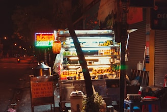 A brightly lit street food stall at night displays an assortment of snacks and beverages. Neon signs, including one with the words 'Bánh Mì', contribute to the vibrant atmosphere. A large pot and other cooking items are visible around the stall, which is situated on a quiet street with minimal traffic.